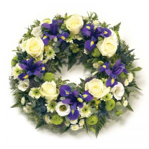 Begravningskrans med rosor, iris, krysantemum och säsongsgrönt. BLommorna går i vitt och lila. Beställa kransen online hos Euroflorist och låt dem buda ut den direkt till platsen för begravningen.